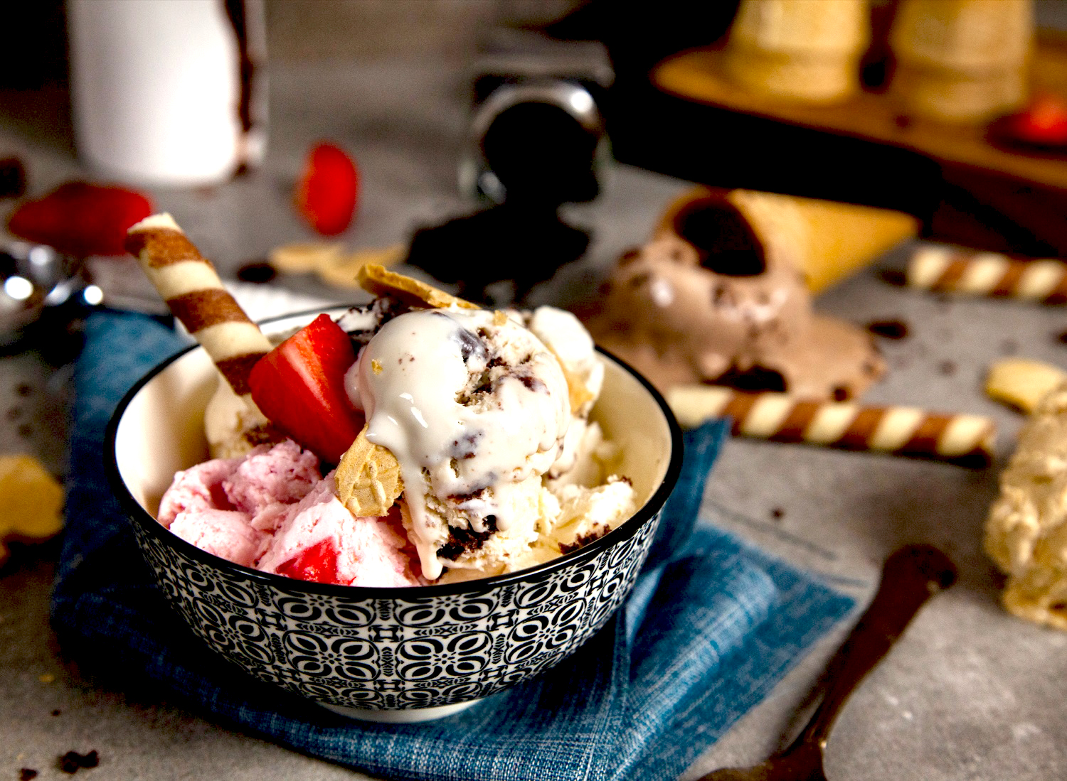 Μπολ σε λευκό και μαύρο χρώμα, επάνω σε τραπέζι με τρούφες σοκολάτας και χωνάκια παγωτού, με σπιτικό παγωτό σε γεύσεις μπισκότο, φράουλα, σοκολάτα, φρέσκες φράουλες και μπισκότα από βανίλια και σοκολάτα, επάνω σε μπλε πετσέτα σερβιρίσματος, με μεταλλικό κουτάλι στο πλάι